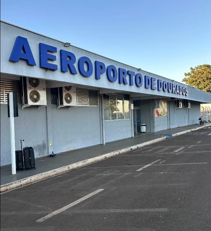 Fachada do terminal do Aeroporto de Dourados, com o letreiro azul Aeroporto de Dourados sobre a marquise e bagagens no passeio