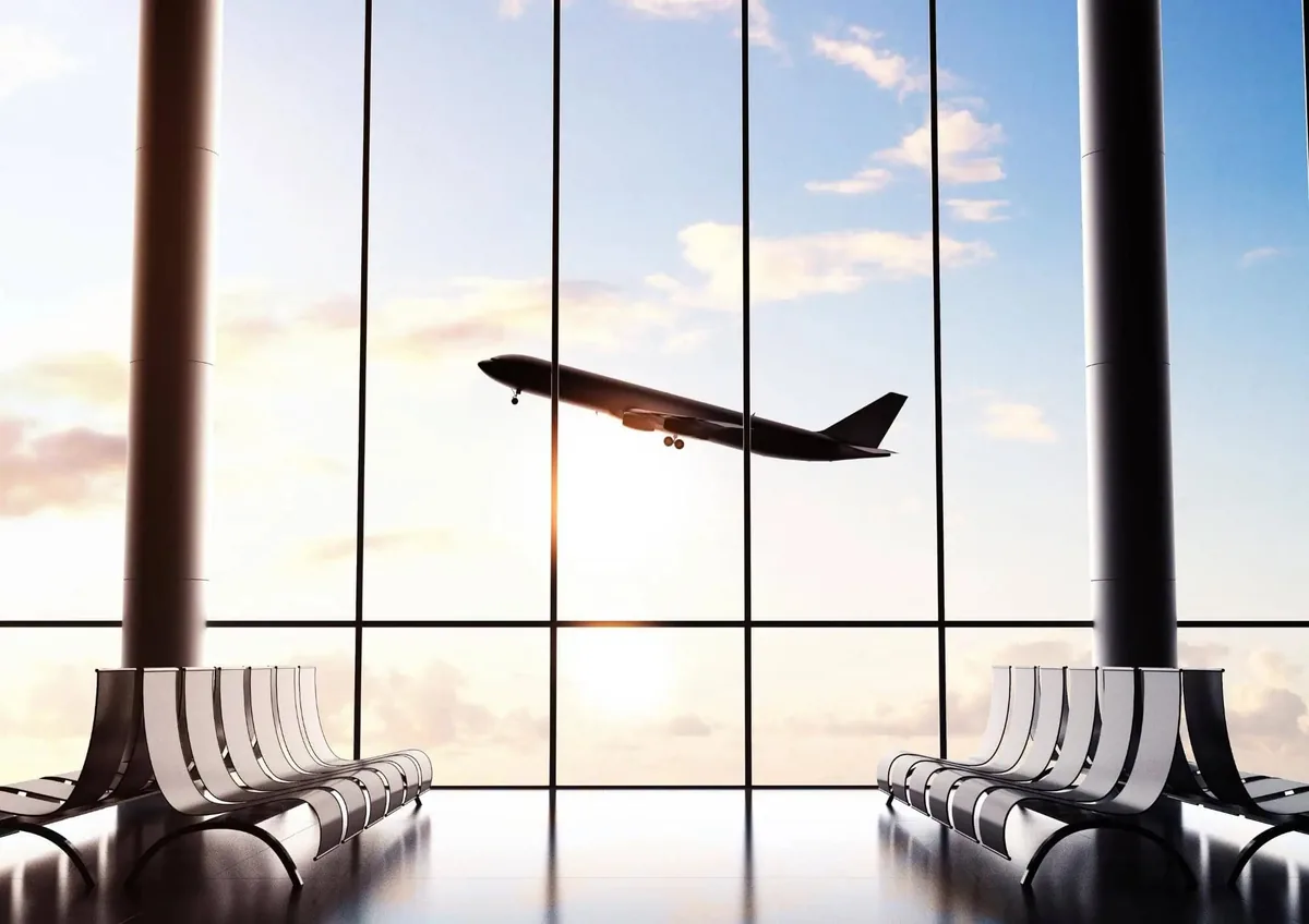 Passageira caminhando pelo terminal com mala de mão enquanto um avião decola ao fundo
