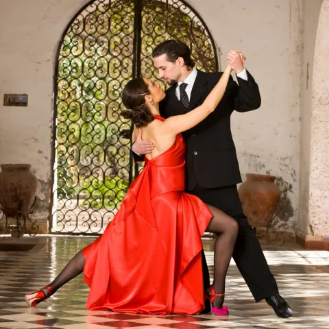 Casal dançando tango, ela de vestido vermelho, em salão