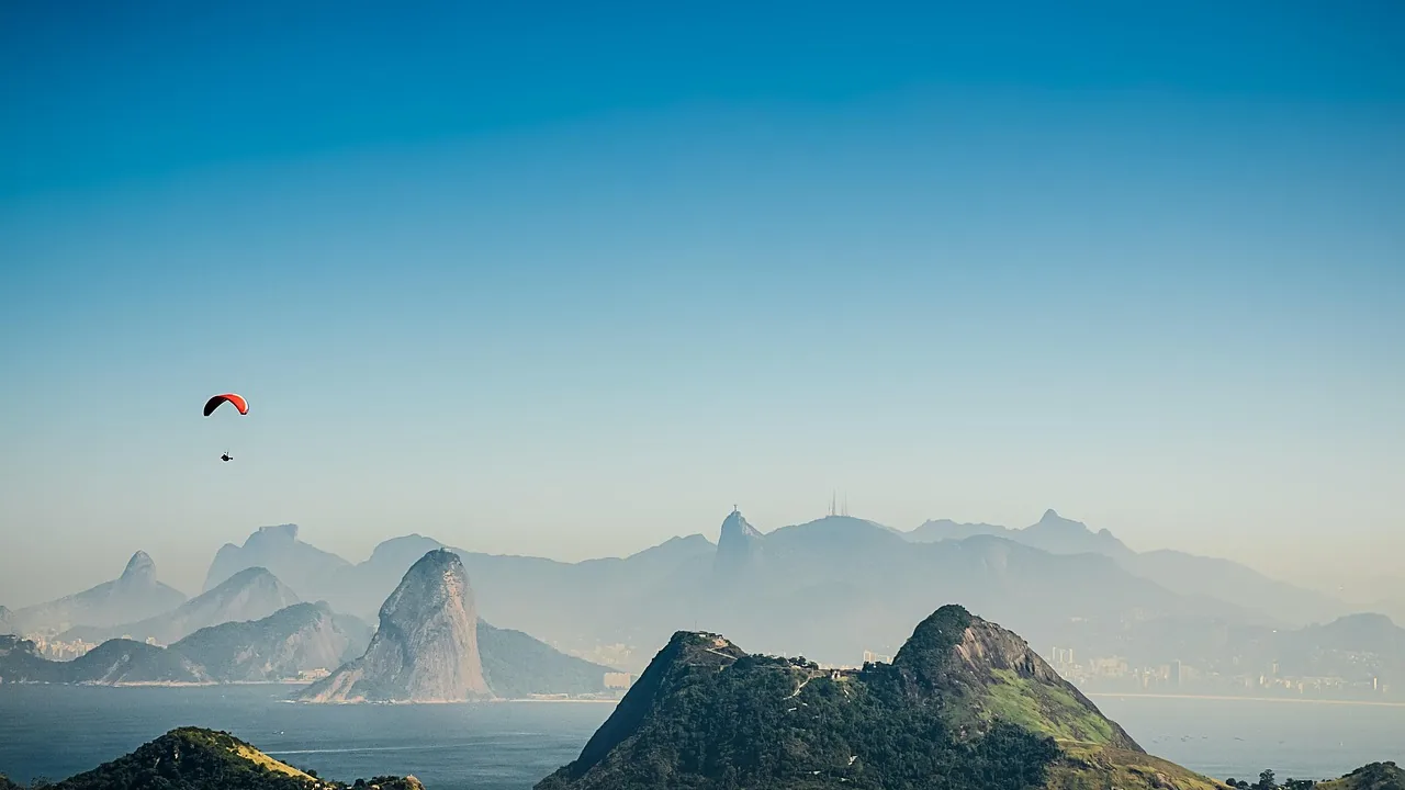 Baía de Guanabara vista de longe, com o Pão de Açúcar e o Corcovado ao fundo e um parapente no céu